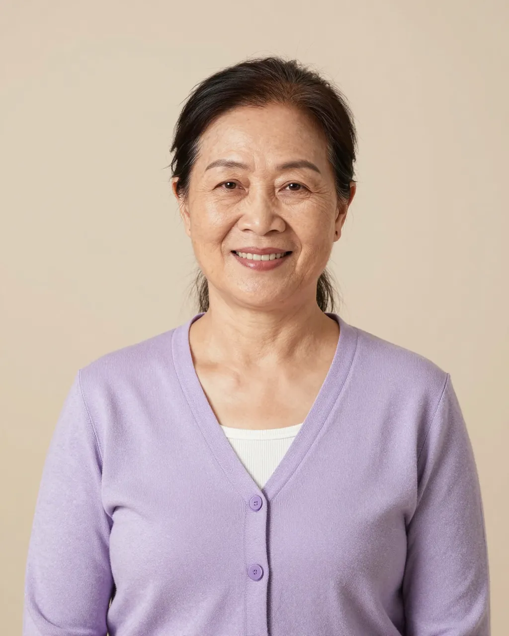 Studio portrait of a smiling senior Southeast Asian woman in a lavender cardigan against a beige backdrop
