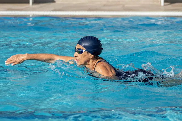 Side view of an older woman in a navy swim cap swimming laps in a bright outdoor pool under morning light.