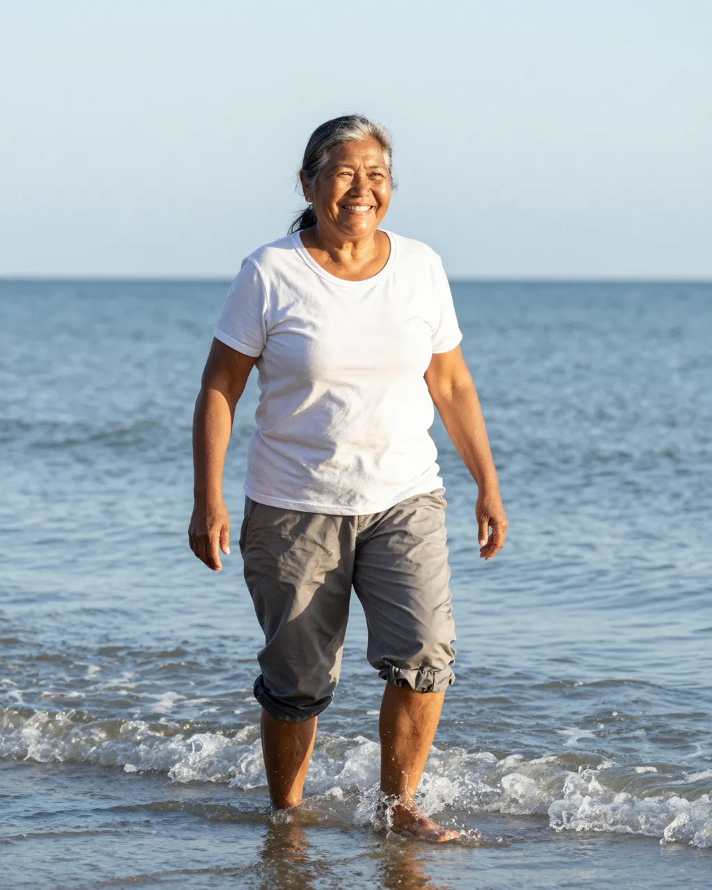 Smiling older woman stands ankle-deep in calm sea water on a bright clear morning