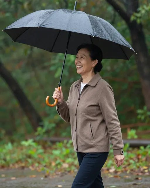 Laughing East Asian woman in her fifties walks with an open umbrella through a forest in warm rain