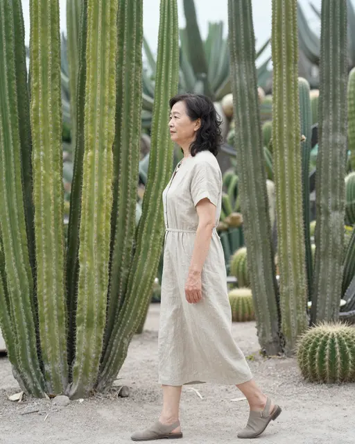 Side view of an East Asian senior woman in a linen dress walking between tall columnar cacti in a desert garden