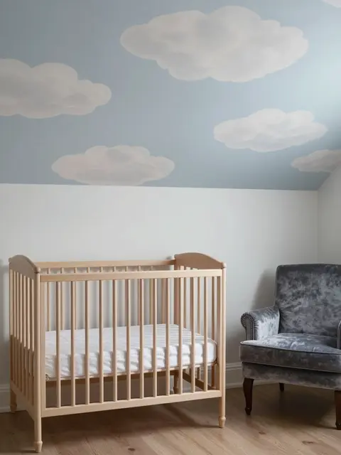 Light wood crib and gray velvet armchair sit under a ceiling painted with soft cloud illustrations