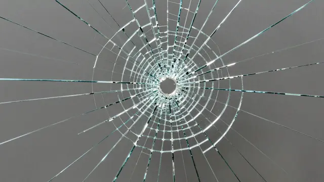 Close-up full frame view of bullet-pattern shattered safety glass with a dense radial crack web against a smooth gray