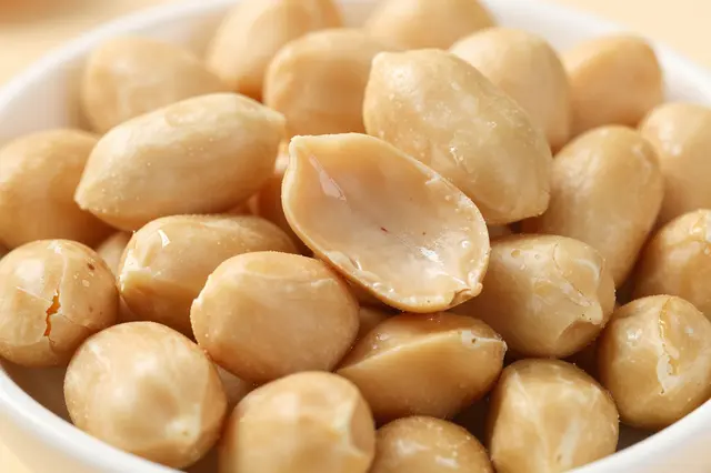 Close-up of shelled roasted peanuts arranged in a small white bowl, with one split peanut in the foreground.