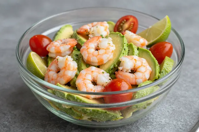 A clear glass bowl holds a fresh salad with cooked shrimp, avocado slices, cherry tomatoes and lime wedges on a gray surface.