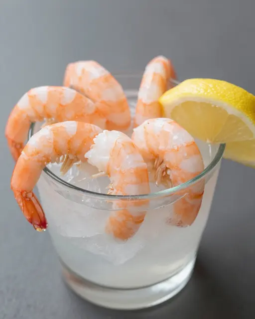 A glass of ice holds cooked shrimp garnished with a fresh lemon wedge on a dark gray surface