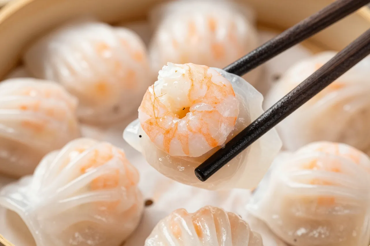Dark chopsticks hold a translucent shrimp dumpling above a bamboo steamer filled with har gow.