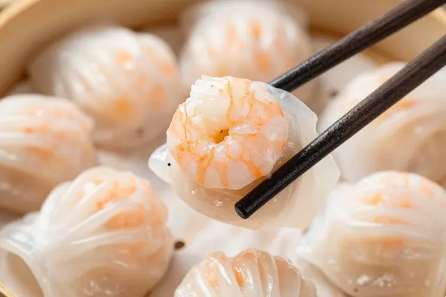 Dark chopsticks hold a translucent shrimp dumpling above a bamboo steamer filled with har gow.