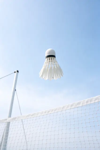 A white badminton shuttlecock hangs in mid-air above a badminton net against a pale blue sky