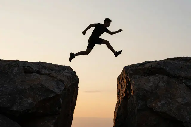 A person's silhouette leaps across a gap between two rocky outcrops at sunset