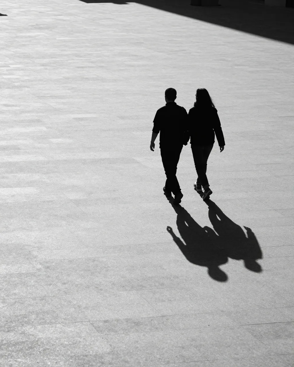Two dark silhouette walkers cast long evening shadows across a pale tiled open plaza