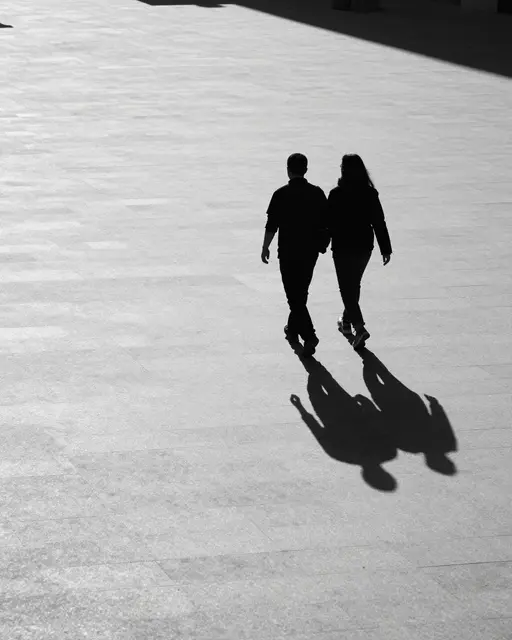 Two dark silhouette walkers cast long evening shadows across a pale tiled open plaza
