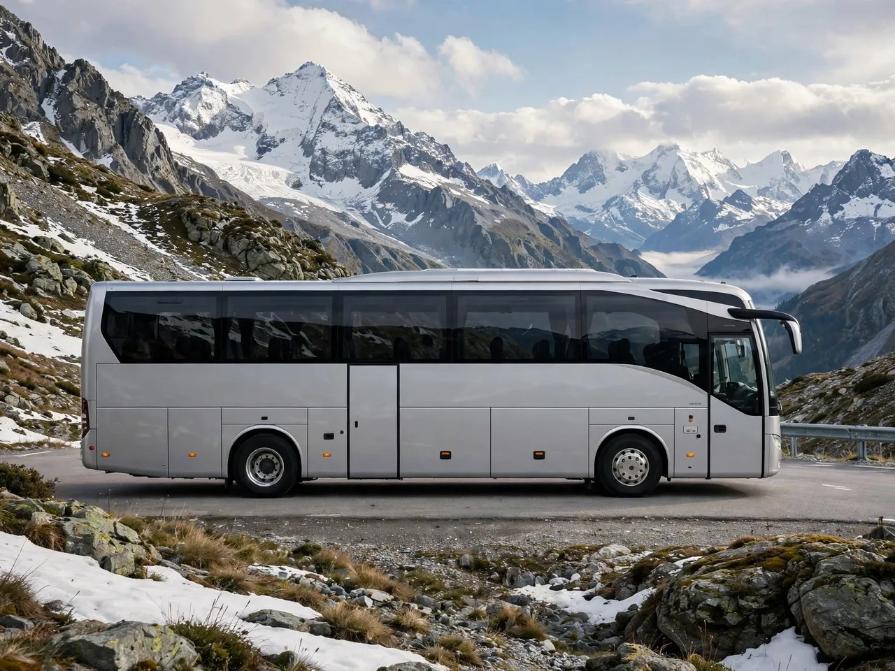 Side view of a plain silver modern coach bus parked on a paved mountain pass road with snow-capped peaks in the background