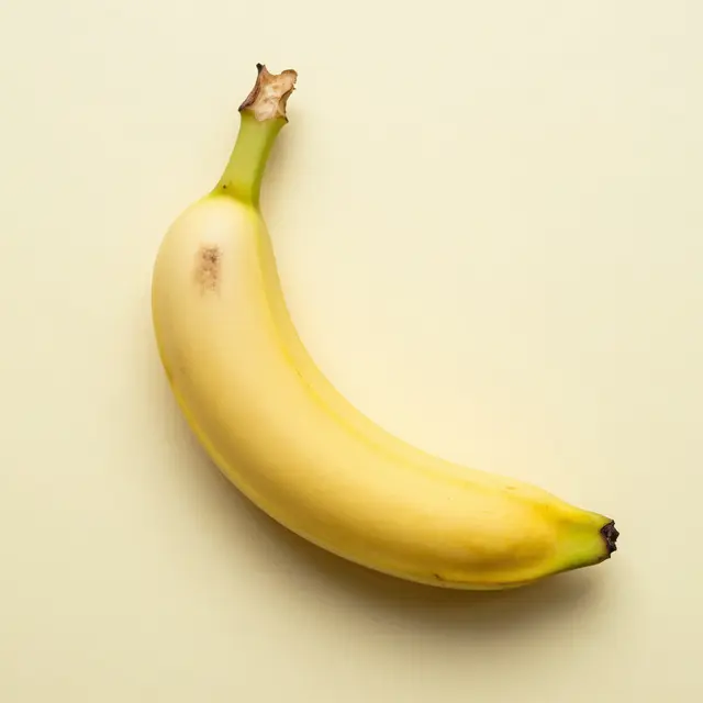 A single ripe yellow banana rests flat on a smooth pastel yellow background