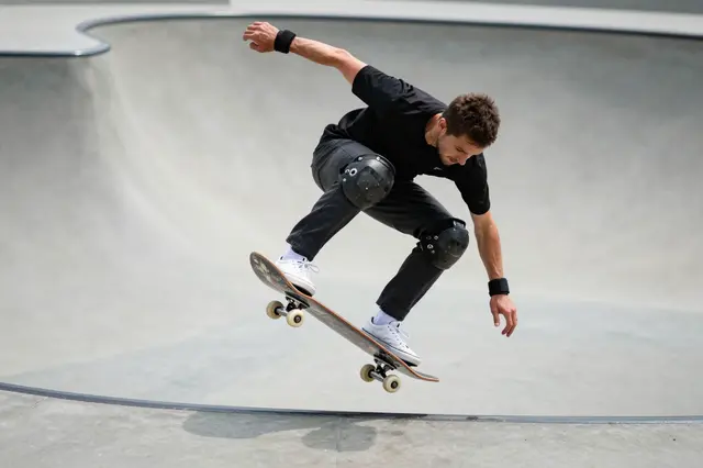 An adult male skateboarder performs an aerial trick above a concrete skatepark bowl edge on a sunny day.