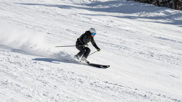 An adult skier in dark gear carves a turn, kicking up a spray of snow on a sunny groomed ski slope.
