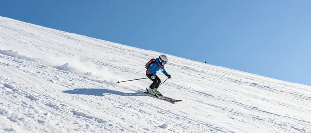 A helmeted skier carves downhill across a vast groomed snowy slope under a clear bright blue sky.