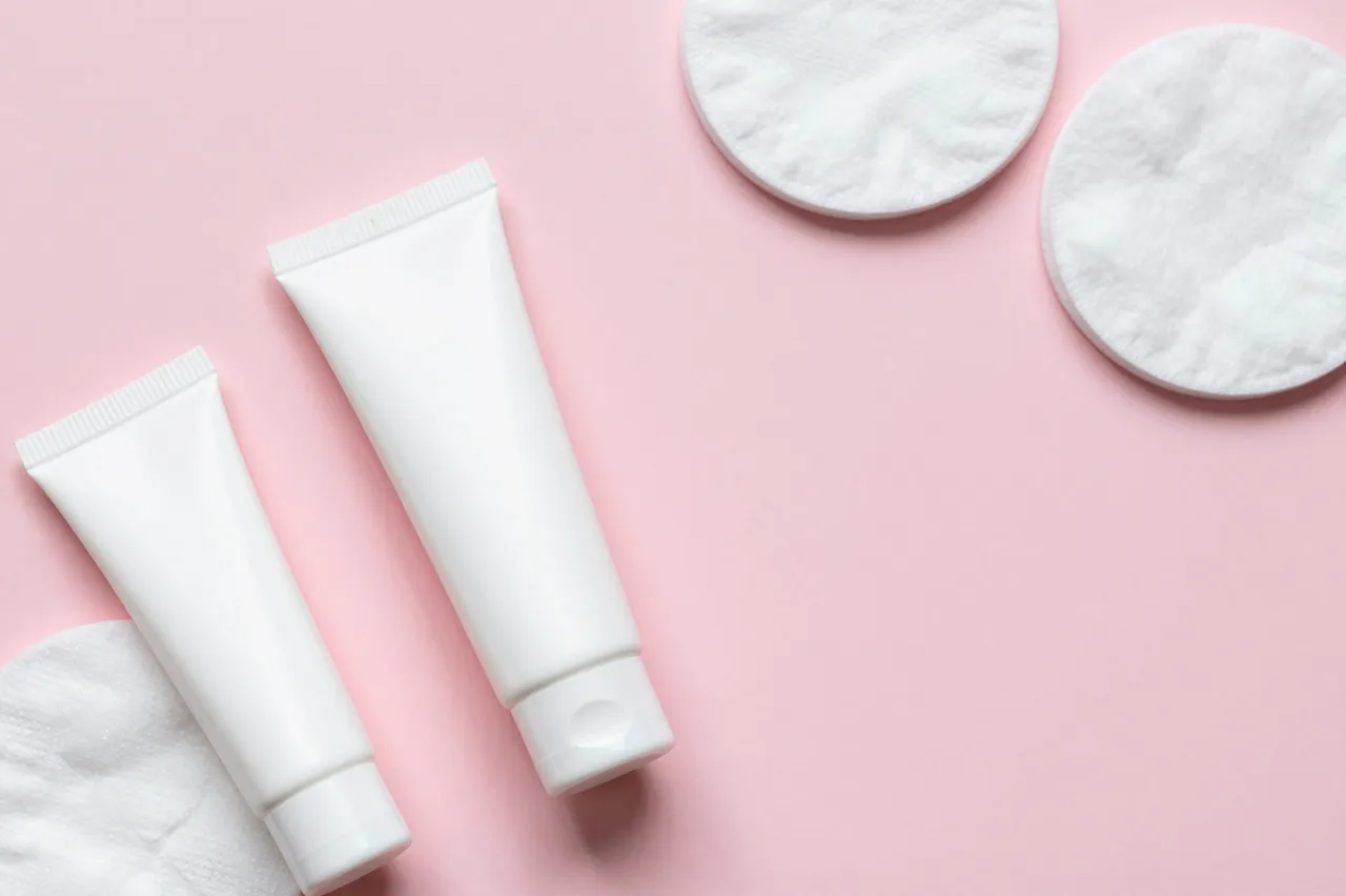 Plain white cosmetic tubes and cotton pads arranged on a blush pink background with empty copy space