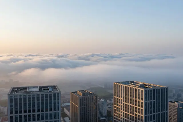 Tall skyscraper rooftops rise above a thick sea of clouds during soft golden sunrise