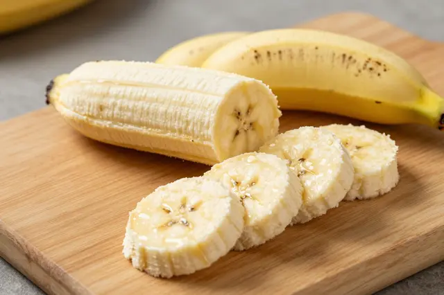 Peeled and sliced banana rests on a wooden cutting board with unpeeled bananas in the background.