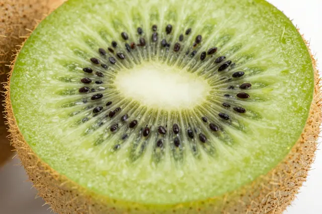 Close up macro shot of a cut kiwi showing its green seeded flesh, fuzzy brown skin, and glistening moisture droplets.