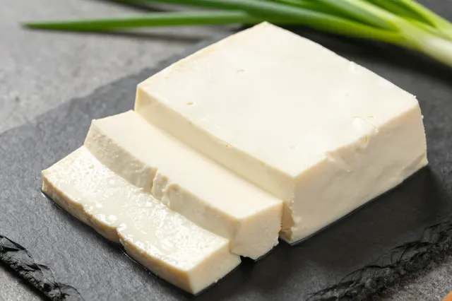 Block of fresh sliced tofu rests on a dark slate with green scallions in the background.