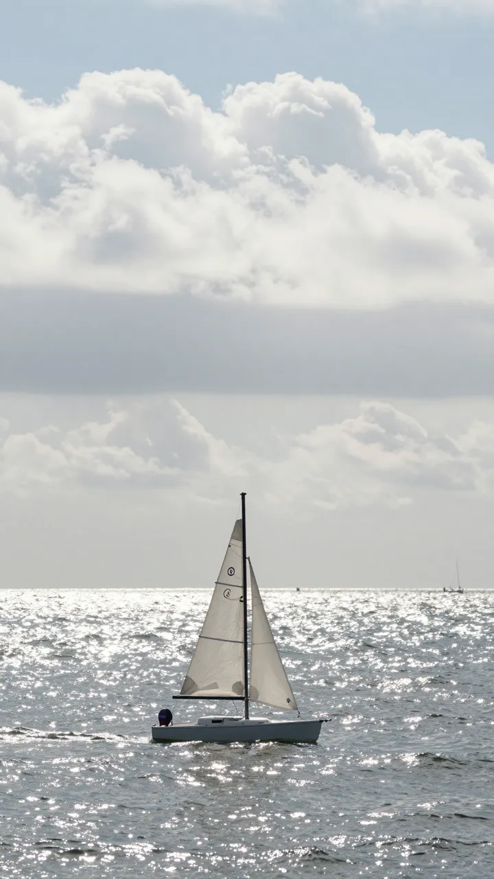 A tiny sailboat floats on a sparkling silver sea beneath large fluffy white clouds on a bright daytime.