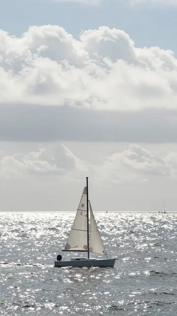 A tiny sailboat floats on a sparkling silver sea beneath large fluffy white clouds on a bright daytime.