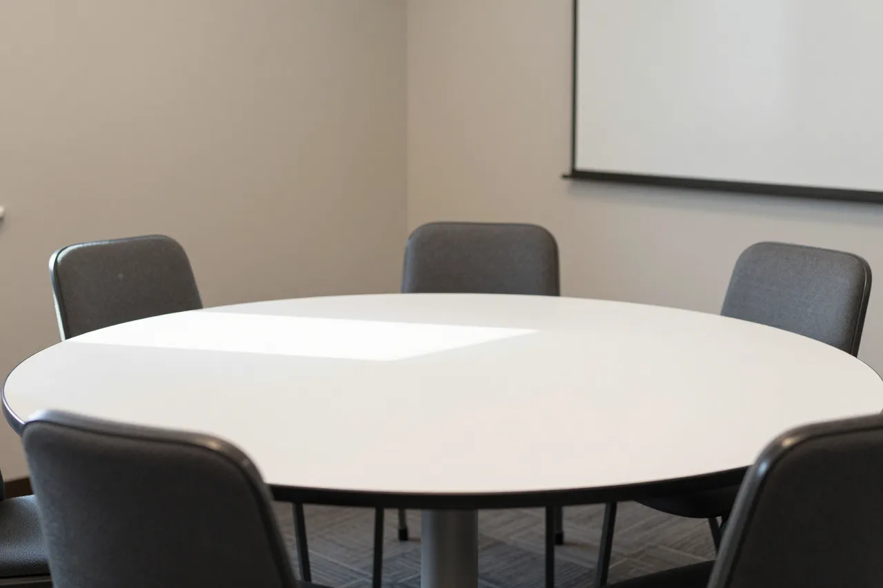 Empty small meeting room with a white round table, gray chairs, and projection screen in soft daylight