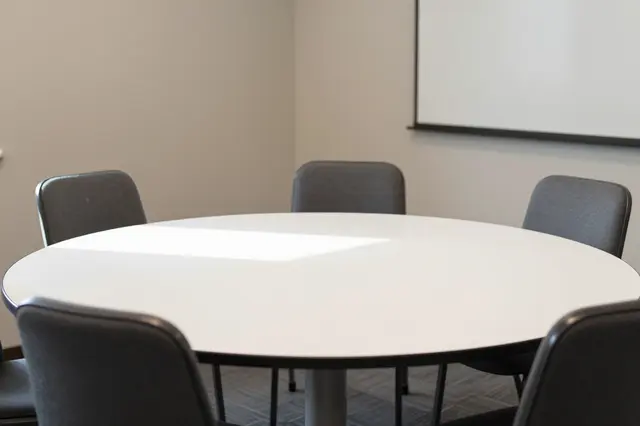 Empty small meeting room with a white round table, gray chairs, and projection screen in soft daylight