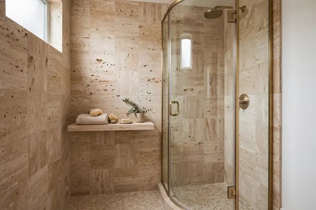 Corner shower with curved brass-framed glass door next to a floating stone shelf on travertine tile walls