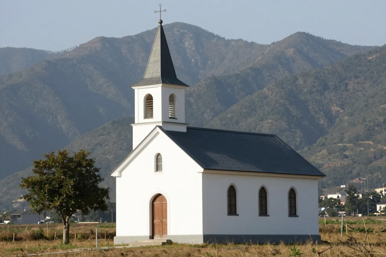 A small white countryside church with a steeple stands in an open field backed by forested mountains