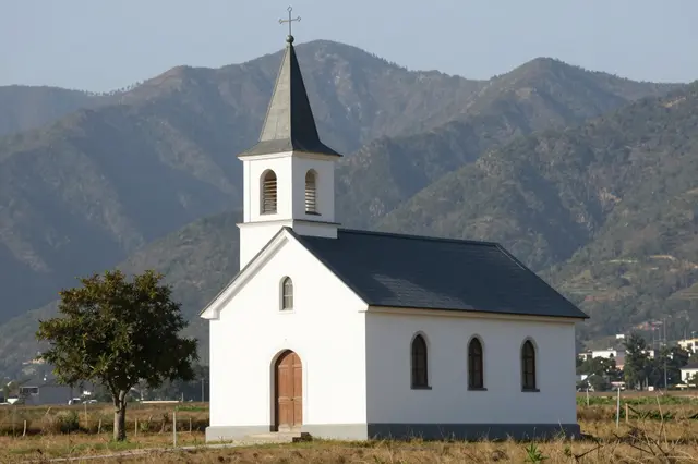 A small white countryside church with a steeple stands in an open field backed by forested mountains