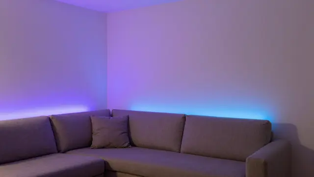 A gray sectional couch sits under walls lit with soft purple and blue smart LED lighting at dusk