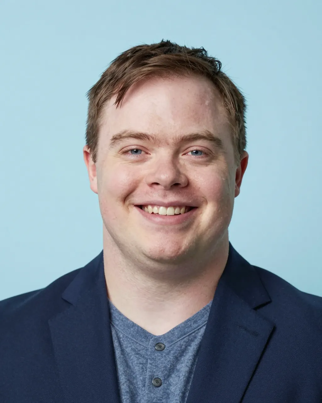 Headshot of a smiling man in his thirties with Down syndrome wearing a navy blazer against a light blue backdrop