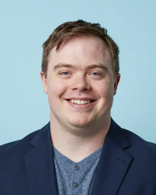Headshot of a smiling man in his thirties with Down syndrome wearing a navy blazer against a light blue backdrop