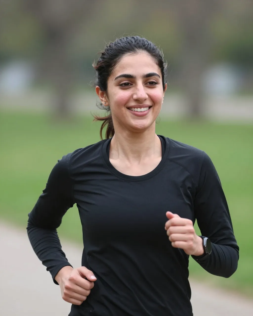 A smiling flushed Middle Eastern woman in athletic wear jogs along a paved park path.