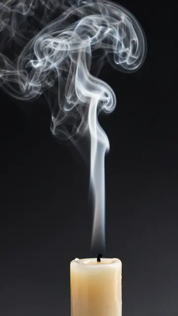 Delicate curling wispy smoke rises from the wick of an extinguished pale candle against a dark black background.