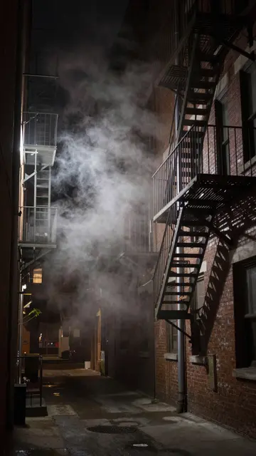 A narrow brick city alley lined with metal fire escapes as steam drifts through the dark night air