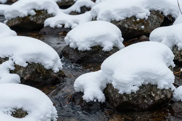 Rounded thick snow caps sit atop dark mossy rocks in a flowing winter stream