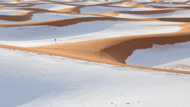 Smooth orange desert dune crests rise above white snow-filled hollows in a rare post-snowfall desert landscape at sunrise