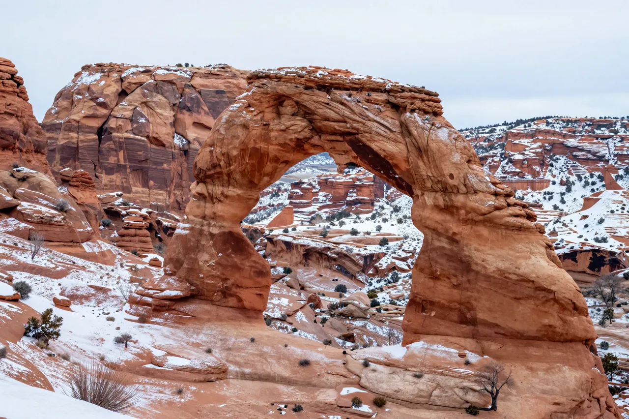 A natural sandstone arch dusted with snow stands over a snow-covered Utah canyon under an overcast winter sky