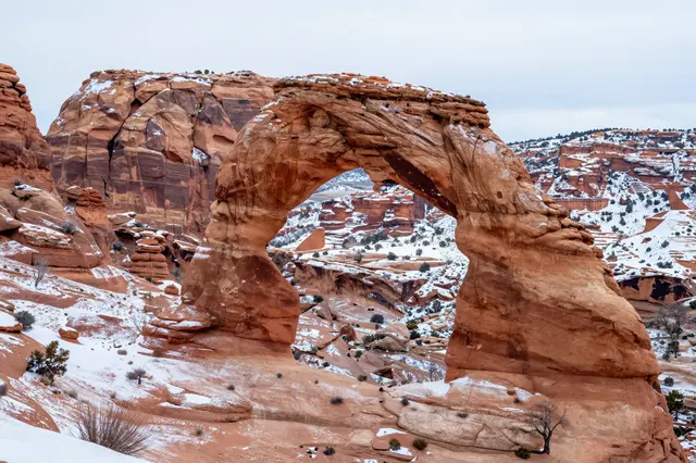 A natural sandstone arch dusted with snow stands over a snow-covered Utah canyon under an overcast winter sky