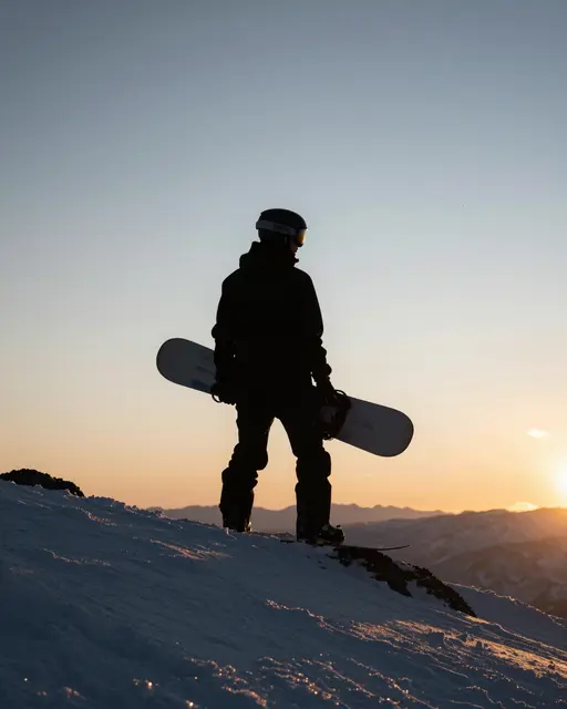 Silhouetted adult snowboarder holding a snowboard stands on a snowy mountain ridge at golden sunset