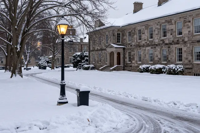 Lit vintage lamp post stands along a snow covered drive beside stone campus buildings at overcast dusk