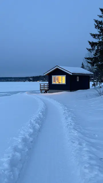 A dug snow path leads to a dark lakeside cabin with warm glowing windows under blue twilight skies