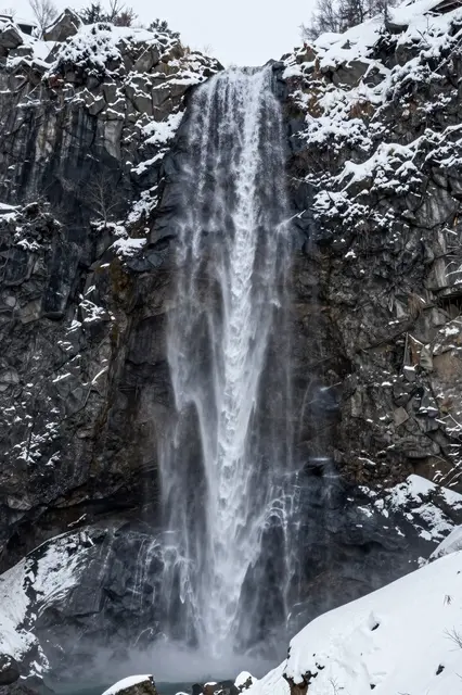 A narrow cascading waterfall drops down a dark snow-dusted rocky mountain cliff in winter