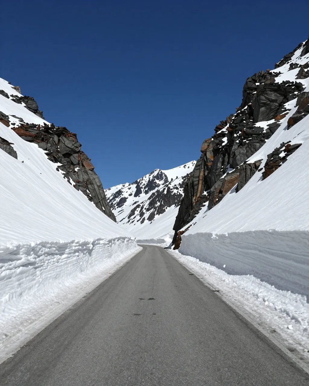 Paved mountain pass road cuts between tall plowed snow banks under a deep clear blue sky