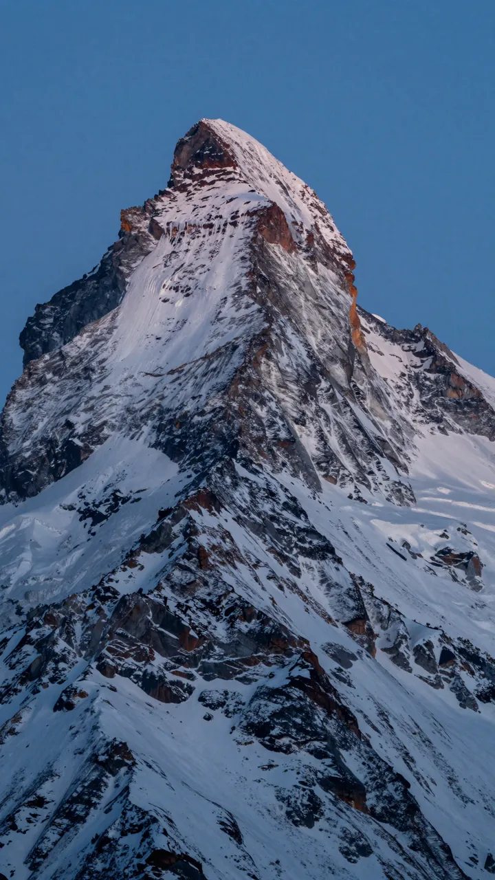 Sharp ridged snowy alpine mountain peak rises against a clear deep blue hour sky