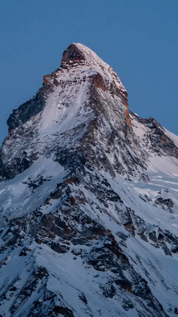 Sharp ridged snowy alpine mountain peak rises against a clear deep blue hour sky